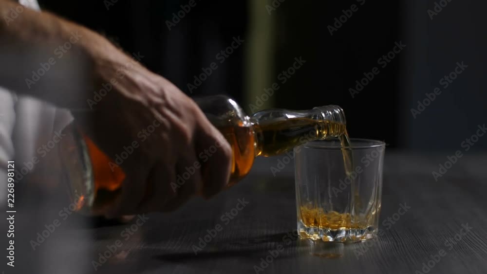 Closeup of male hand holding bottle of alcohol beverage and refilling glass with whisky. Drunk wasted male alcoholic with shaking hands in alcohol abuse pouring whiskey into glass at home in evening.