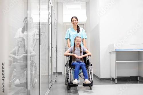 Young female doctor taking care of little girl in wheelchair indoors