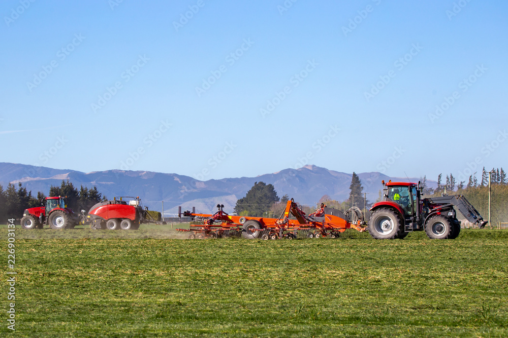 Fototapeta premium Red farm machinery at work in a rural field in Canterbury, New Zealand, raking and baling hay