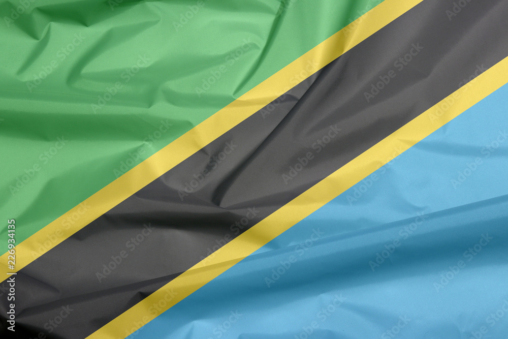 Fabric flag of Tanzania. Crease of Tanzanian flag background, A yellow-edged black diagonal band: the upper triangle is green and the lower triangle is blue. Fabric flag of Tanzania. Crease of Tanzanian flag background, A yellow-edged black diagonal band: the upper triangle is green and the lower triangle is blue.
