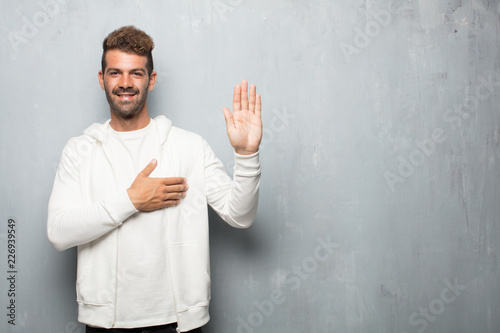 Принти на полотні young handsome man smiling confidently while making a sincere promise or oath, solemnly swearing with one hand over heart