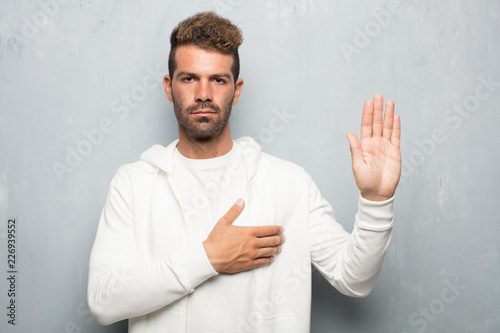фотографія young handsome man smiling confidently while making a sincere promise or oath, solemnly swearing with one hand over heart