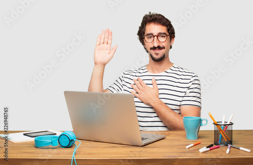 фотографія young bearded graphic designer with a laptop smiling confidently while making a sincere promise or oath, solemnly swearing with one hand over heart