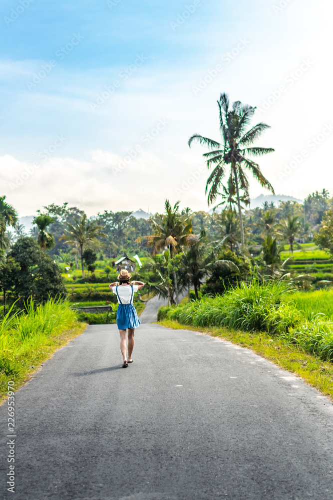 Tropical portrait of young happy woman with straw hat on a road with coconut palms and tropical trees. Bali island.