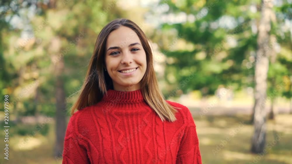 Slow motion portrait of beautiful young woman in bright red sweater standing in the park with happy smile and looking at camera. Nature and youth concept.