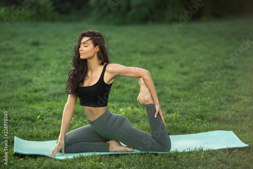 Woman practicing yoga performing yoga-asanas. Young attractive slim girl in bodysuit relaxing and doing exercises, sitting in variation of One Legged King Pigeon Posture on a yoga mat. Gorgeous