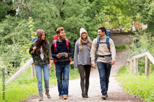 travel, technology and hiking concept - group of smiling friends or travelers with backpacks and tablet pc computers walking along road