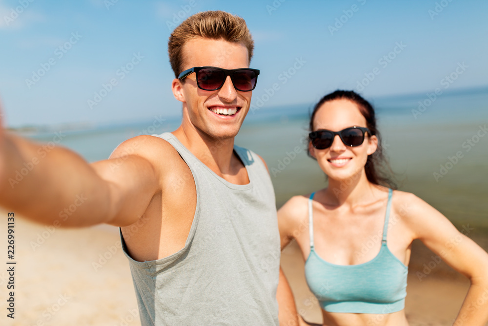 fitness, sport and lifestyle concept - happy couple in sports clothes taking selfie by smartphone on beach