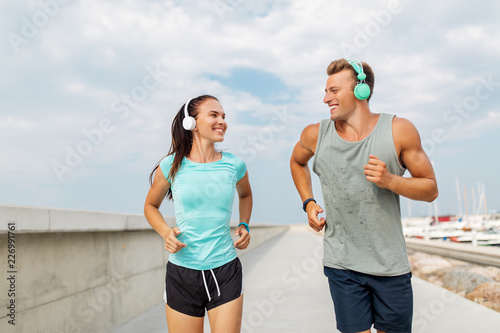 sport  people and technology concept - happy couple with headphones and fitness trackers running outdoors