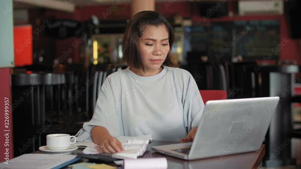 Young concentrated Asian woman sitting at restaurant table, looking at receipts and working on laptop while calculating month profit Young concentrated Asian woman sitting at restaurant table, looking at receipts and working on laptop while calculating month profit