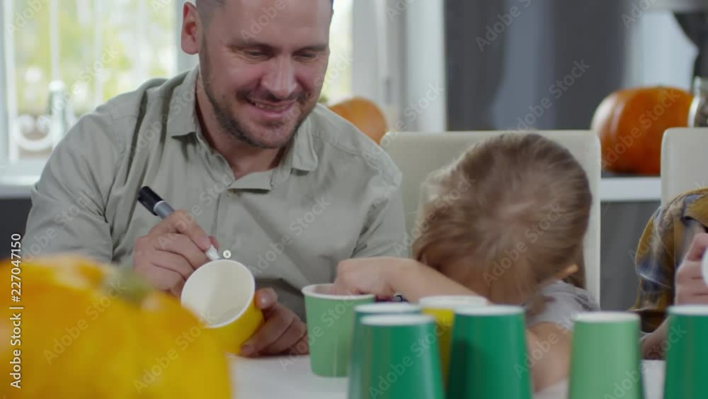 Joyous father smiling and drawing on paper cup with felt tip pen while preparing Halloween decorations with little daughter Joyous father smiling and drawing on paper cup with felt tip pen while preparing Halloween decorations with little daughter