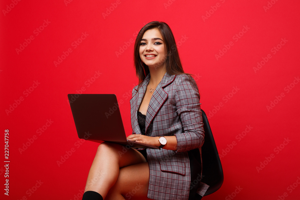 Young cheerful girl sitting on a chair and holding a laptop on his feet.Young student wearing a striped jacket.Red background Young cheerful girl sitting on a chair and holding a laptop on his feet.Young student wearing a striped jacket.Red background
