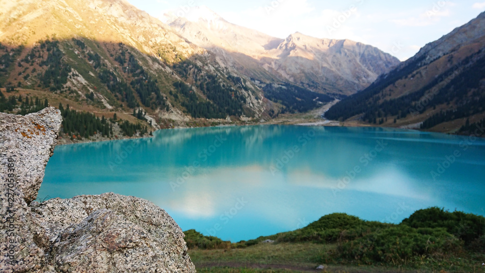 Beautiful view of the mountain lake of heavenly color. The color of the water reflected the oboe and the mountains. On the sides the grasses, coniferous trees, ate. Visible stones in the foreground.