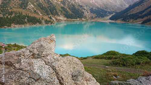Beautiful view of the mountain lake of heavenly color. The color of the water reflected the oboe and the mountains. On the sides the grasses  coniferous trees  ate. Visible stones in the foreground.