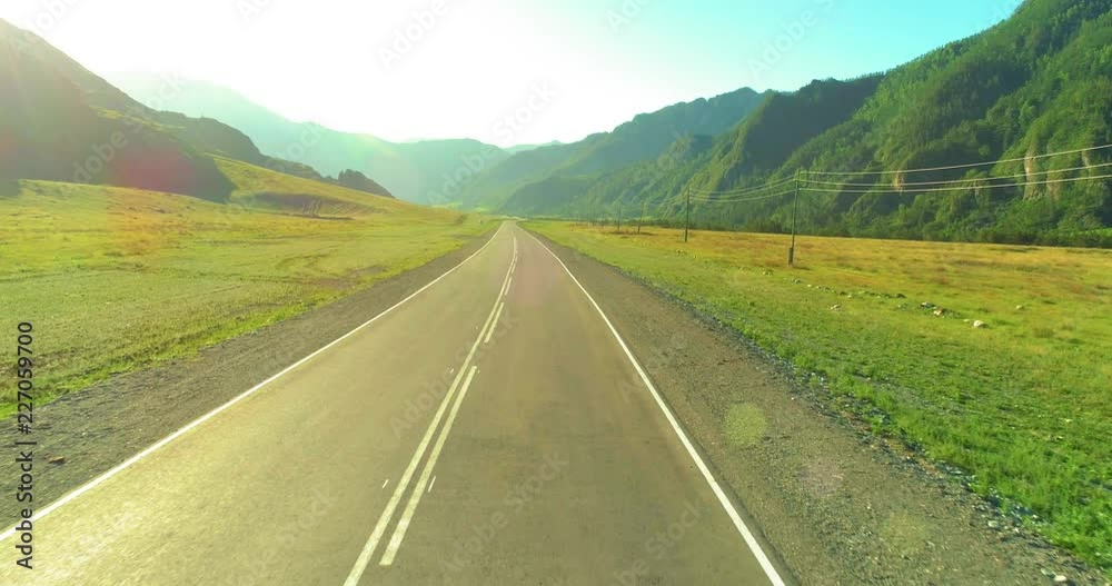 Aerial low air flight over mountain perfect asphalt highway road and meadow at sunny summer morning. Near green trees. Fast horizontal movement with sun rays. Aerial low air flight over mountain perfect asphalt highway road and meadow at sunny summer morning. Near green trees. Fast horizontal movement with sun rays.