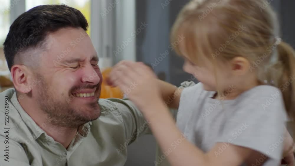 Close-up of happy father sitting with little daughter at home and laughing while she playing with his nose
