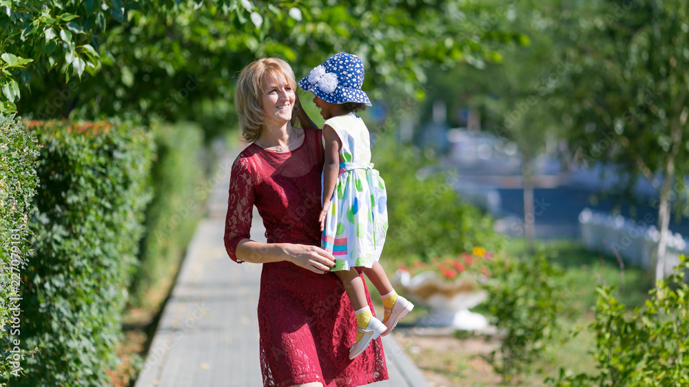 A white woman carries a black girl on her shoulder. European mom and dark-skinned foster daughter have fun on a walk. A white woman carries a black girl on her shoulder. European mom and dark-skinned foster daughter have fun on a walk.