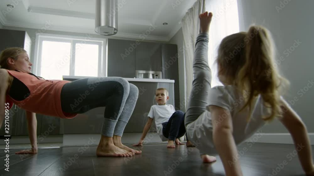 Medium shot of young mother in sportswear showing exercise to little children repeating after her: she is standing in reverse tabletop pose and doing leg kicks Medium shot of young mother in sportswear showing exercise to little children repeating after her: she is standing in reverse tabletop pose and doing leg kicks