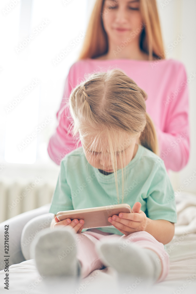 Young mother styling daughter's hair while she's watching cartoons on smart phone Young mother styling daughter's hair while she's watching cartoons on smart phone