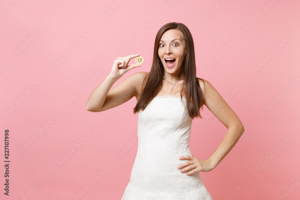 Excited bride woman in white wedding dress holding bitcoin metal coin of golden color future currency isolated on pink pastel background. Organization of wedding concept. Copy space for advertisement. Excited bride woman in white wedding dress holding bitcoin metal coin of golden color future currency isolated on pink pastel background. Organization of wedding concept. Copy space for advertisement.