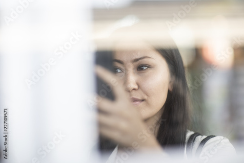 Junge Frau aus Asien schaut auf Smartphone hinter Glasfront