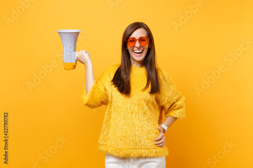 Portrait of cheerful attractive young woman in fur sweater orange heart eyeglasses holding megaphone isolated on bright yellow background. People sincere emotions, lifestyle concept. Advertising area.