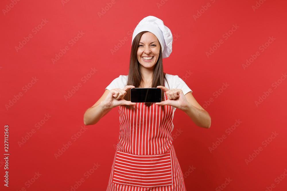 Housewife female chef cook or baker in striped apron white t-shirt toque chefs hat isolated on red wall background. Woman hold cellphone blank screen for promotional content Mock up copy space concept Housewife female chef cook or baker in striped apron white t-shirt toque chefs hat isolated on red wall background. Woman hold cellphone blank screen for promotional content Mock up copy space concept