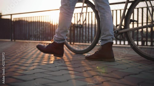 Close up shot of young stylish hipster walking with his bicycle on sea port background during sunset, slow motion photo