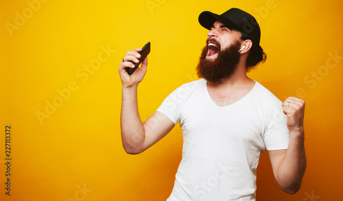 Portrait of happy bearded hipster man singing and listening to music with cell phone and wireless earphones isolated over yellow wall