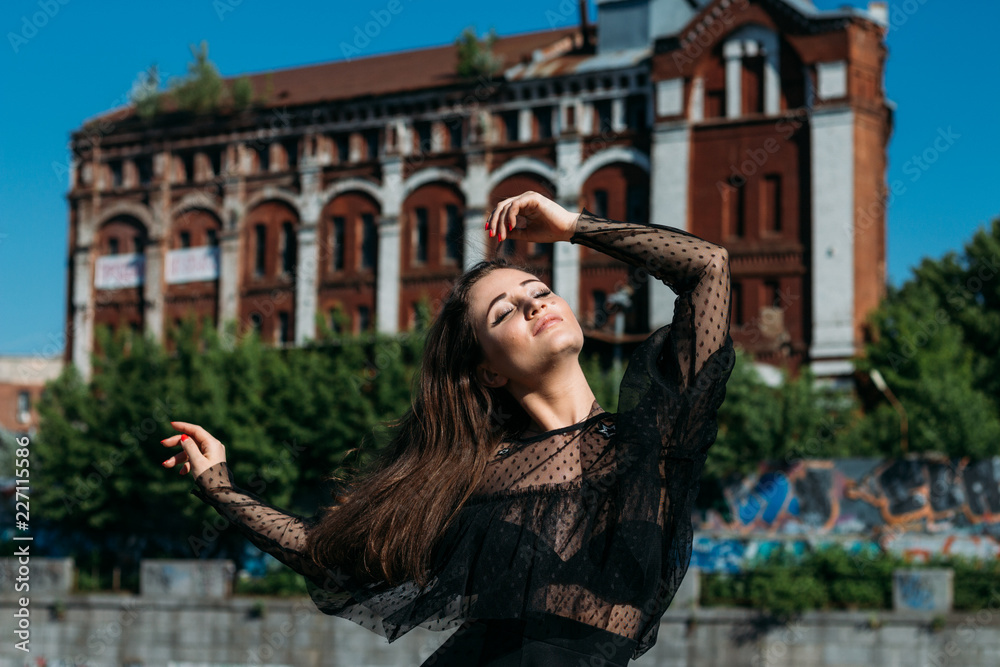 Obraz premium beautiful girl, brunette, in the summer by the river in the city, in a black dress. standing alone by the river, on the bridge, the sun is shining in the face, closed her eyes, rest and tourism.