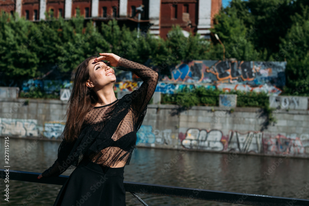 beautiful girl, brunette, in the summer by the river in the city, in a black dress. standing alone by the river, on the bridge, the sun is shining in the face, closed her eyes, rest and tourism.
