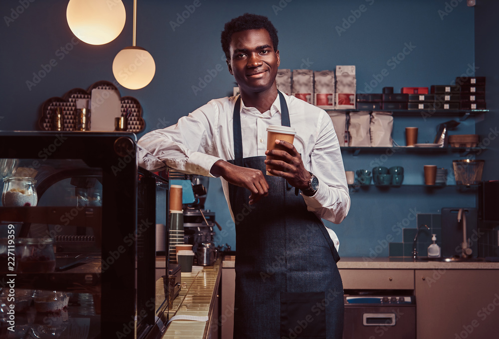African barista smiling at camera relaxing after workday with coffee while leaning on the counter at coffee shop.