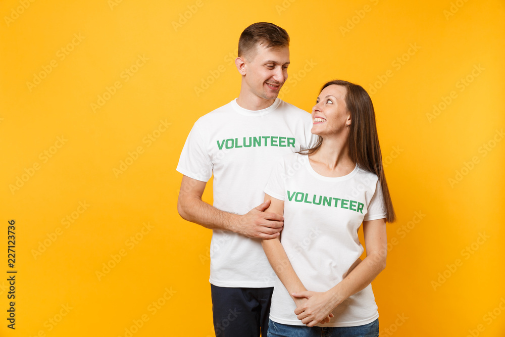 Fototapeta premium Portrait of young colleagues couple in white t-shirt with written inscription green title volunteer isolated on yellow background. Voluntary free work, assistance help, charity grace teamwork concept.