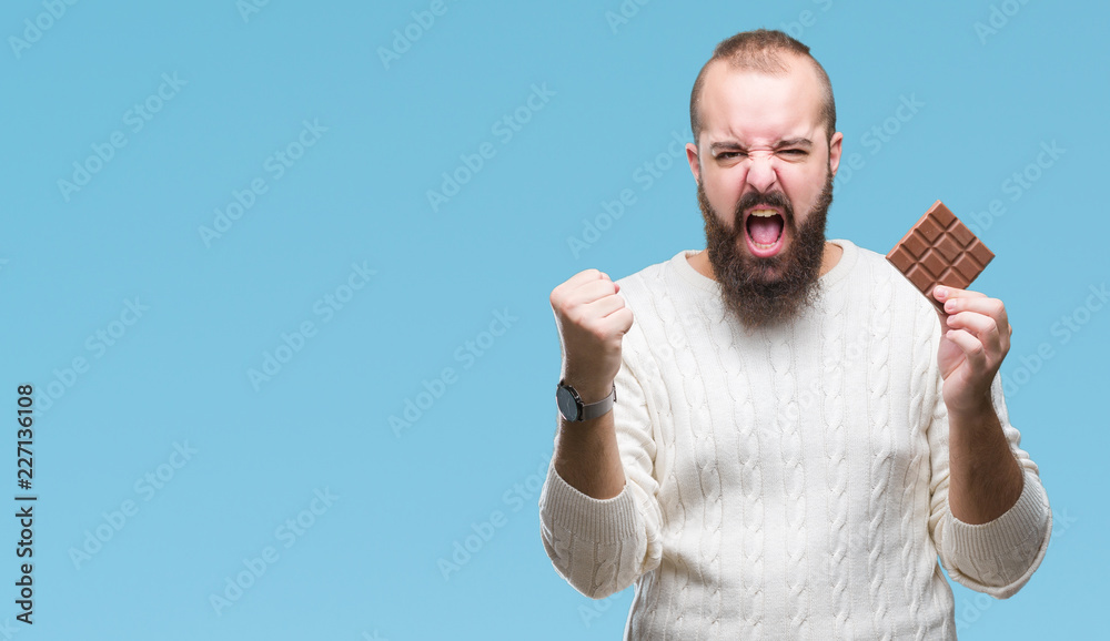 Young hipster man eating chocolate bar over isolated background annoyed and frustrated shouting with anger, crazy and yelling with raised hand, anger concept Young hipster man eating chocolate bar over isolated background annoyed and frustrated shouting with anger, crazy and yelling with raised hand, anger concept