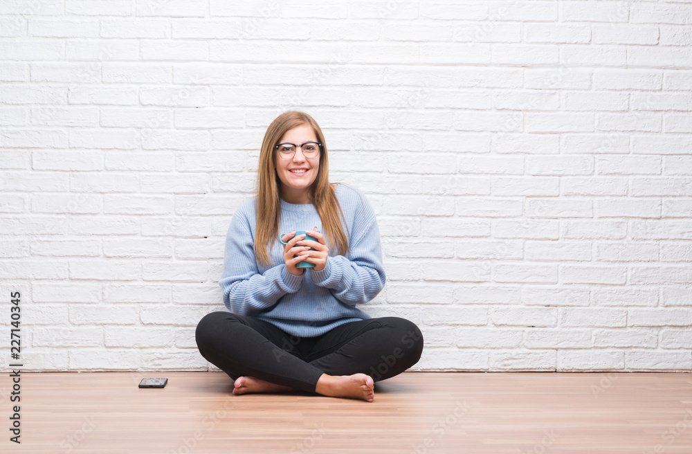 Young adult woman sitting on the floor over white brick wall driking cup of coffee with a happy face standing and smiling with a confident smile showing teeth Young adult woman sitting on the floor over white brick wall driking cup of coffee with a happy face standing and smiling with a confident smile showing teeth
