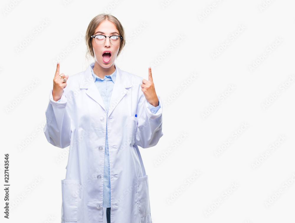 Beautiful young blonde doctor woman wearing white coat over isolated background amazed and surprised looking up and pointing with fingers and raised arms.
