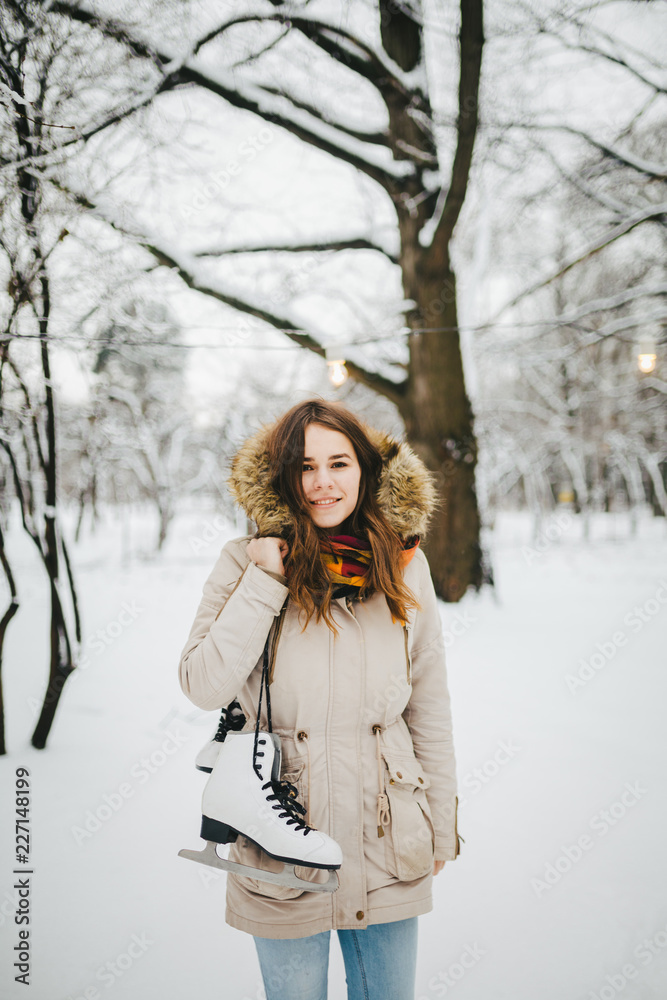 Theme is weekend holiday in winter. A beautiful young Caucasian woman stands in a snow covered park in jacket with hood and fur in jeans and holds on shoulder pair of white skates for long shoelaces