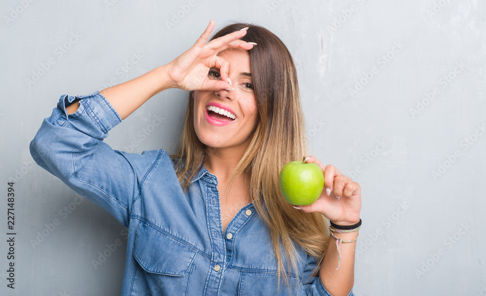 Young adult woman over grey grunge wall eating fresh green apple with happy face smiling doing ok sign with hand on eye looking through fingers Young adult woman over grey grunge wall eating fresh green apple with happy face smiling doing ok sign with hand on eye looking through fingers