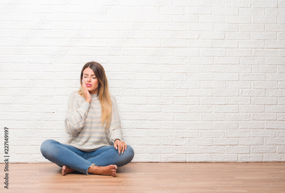 Young adult woman sitting on the floor over white brick wall at home touching mouth with hand with painful expression because of toothache or dental illness on teeth. Dentist concept. Young adult woman sitting on the floor over white brick wall at home touching mouth with hand with painful expression because of toothache or dental illness on teeth. Dentist concept.