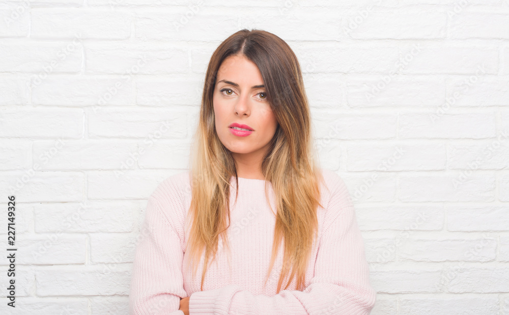 Young adult woman over white brick wall wearing winter outfit at home with serious expression on face. Simple and natural looking at the camera. Young adult woman over white brick wall wearing winter outfit at home with serious expression on face. Simple and natural looking at the camera.