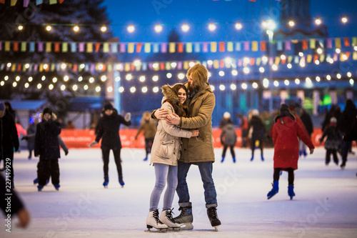Young couple in love Caucasian man with blond hair with long hair and beard and beautiful woman have fun, active date skating on ice scene in town square in winter on Christmas Eve