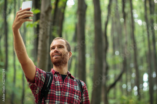Happy young bearded man taking a selfie while hiking through forest on a beautiful autumn day. Healthy and active outdoor lifestyle