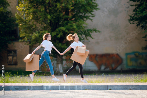 Two young happy female friends in casual clothes with shopping bags running on the street holding hands.