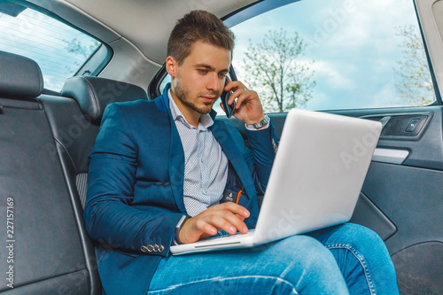 бизнесмен сидит на заднем сиденье авто с ноутбуком, разговаривает по телефону © lufotolab