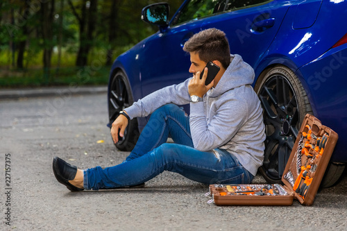 человек сидит возле сломанной машины с инструментами и звонит по телефону, чтобы позвонить в службу