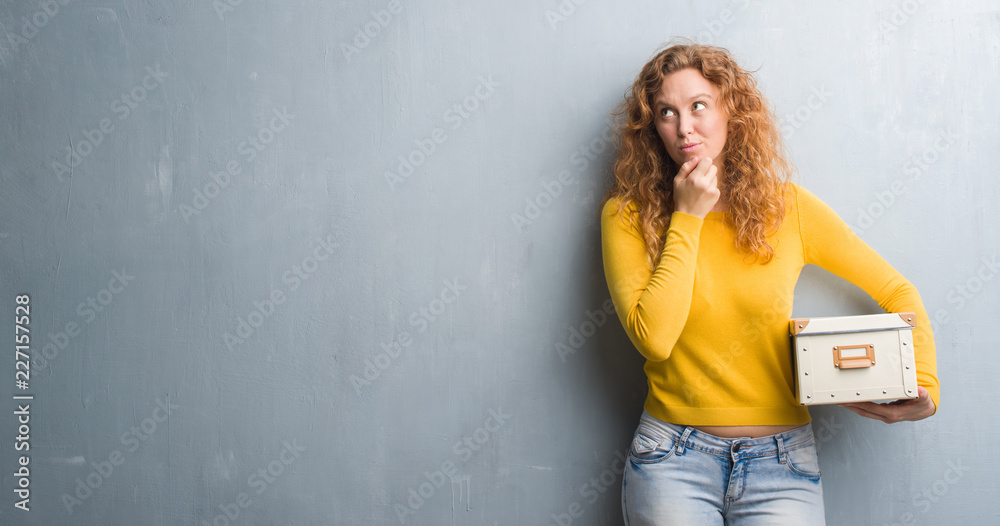 Young redhead woman over grey grunge wall holding box serious face thinking about question, very confused idea Young redhead woman over grey grunge wall holding box serious face thinking about question, very confused idea