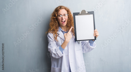 Young redhead doctor woman over grey grunge wall showing clipboard very happy pointing with hand and finger