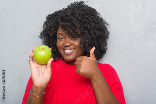 Young african american woman over grey grunge wall eating green apple happy with big smile doing ok sign  thumb up with fingers  excellent sign