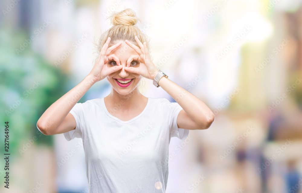 Young beautiful blonde woman wearing white t-shirt over isolated background doing ok gesture like binoculars sticking tongue out, eyes looking through fingers. Crazy expression.