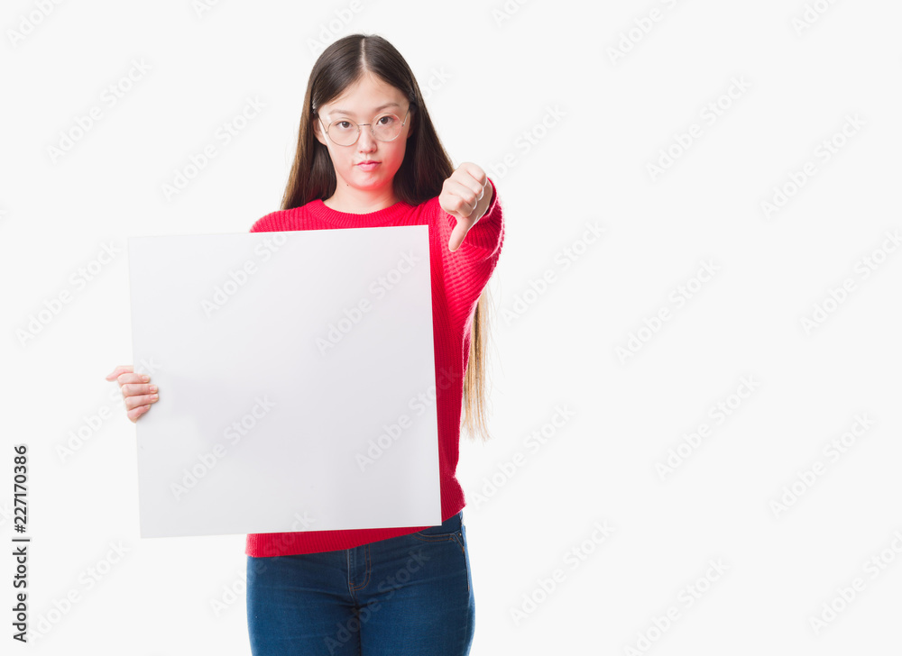 Young Chinese woman over isolated background holding blank banner with angry face, negative sign showing dislike with thumbs down, rejection concept Young Chinese woman over isolated background holding blank banner with angry face, negative sign showing dislike with thumbs down, rejection concept